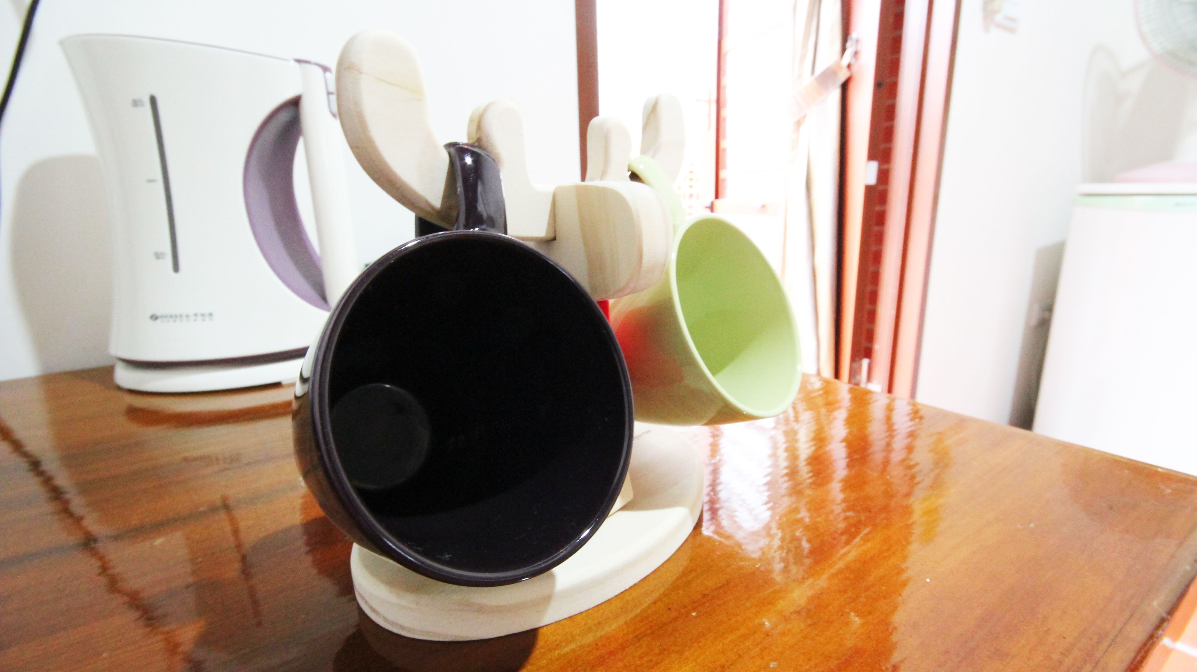 Una zona de cocina con cafetera, una taza negra y un juego de tazas de colores sobre una mesa de madera.