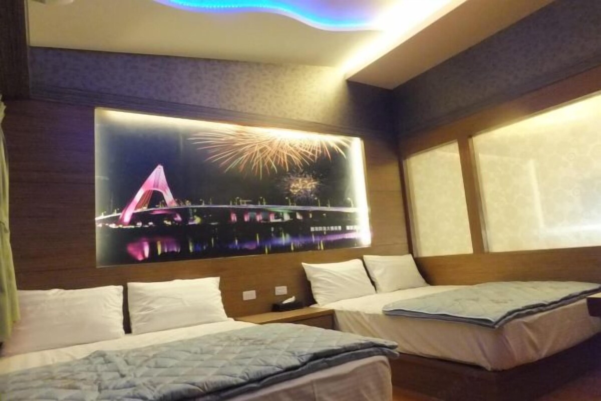 Habitación de hotel con dos camas, un televisor grande montado en la pared mostrando fuegos artificiales y una ventana con vista al cielo nocturno.