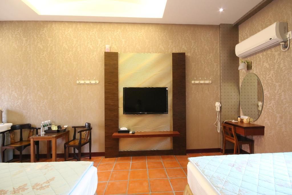 Habitación de hotel con una televisión de pantalla plana montada en una pared texturizada, un escritorio con una silla, una mesita de noche y una cama con cubrecama acolchado.