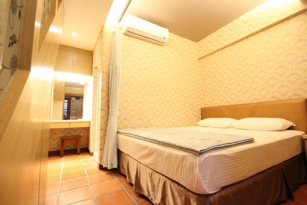 Un dormitorio con cama, mesita de noche, unidad de aire acondicionado y un taburete pequeño.