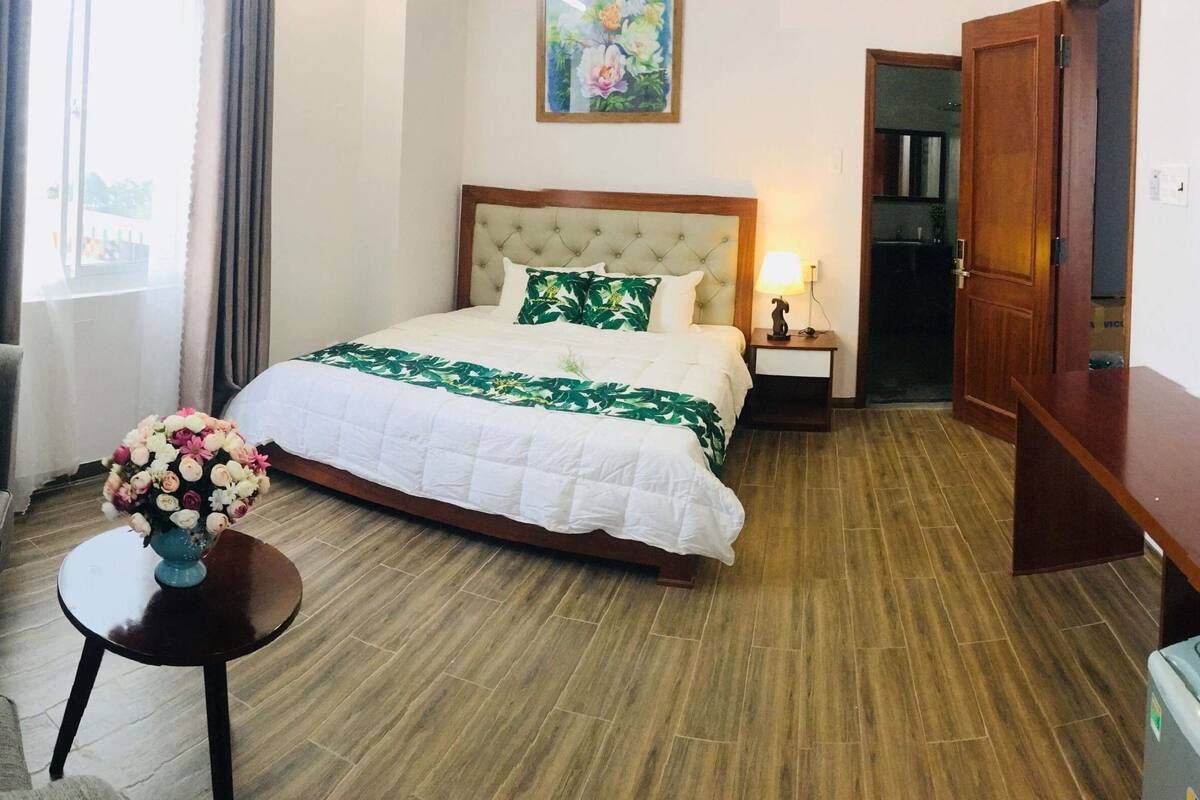 A hotel room with a bed, a sofa, a small table with a vase of flowers, a nightstand, a TV, and a door.