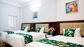 A hotel room with two beds, each with a green leaf-patterned bedspread, white pillows, and a wooden headboard. There is a window with blinds, a vase with flowers on the nightstand, and a painting on the wall.