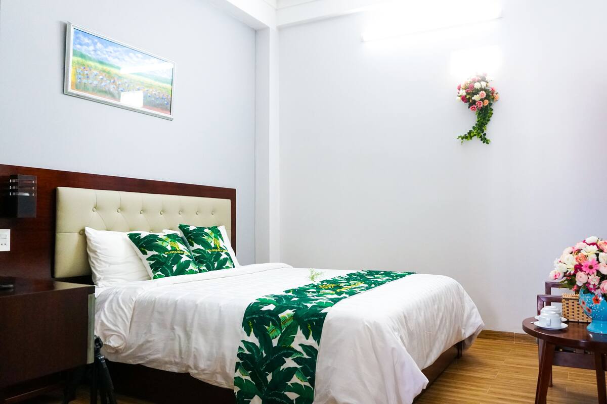A hotel room with a bed, a nightstand, a desk, a chair, a vase with flowers, and a painting on the wall.