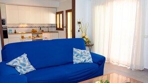 Un salon avec un canapé bleu, une table basse en verre et une cuisine en arrière-plan.