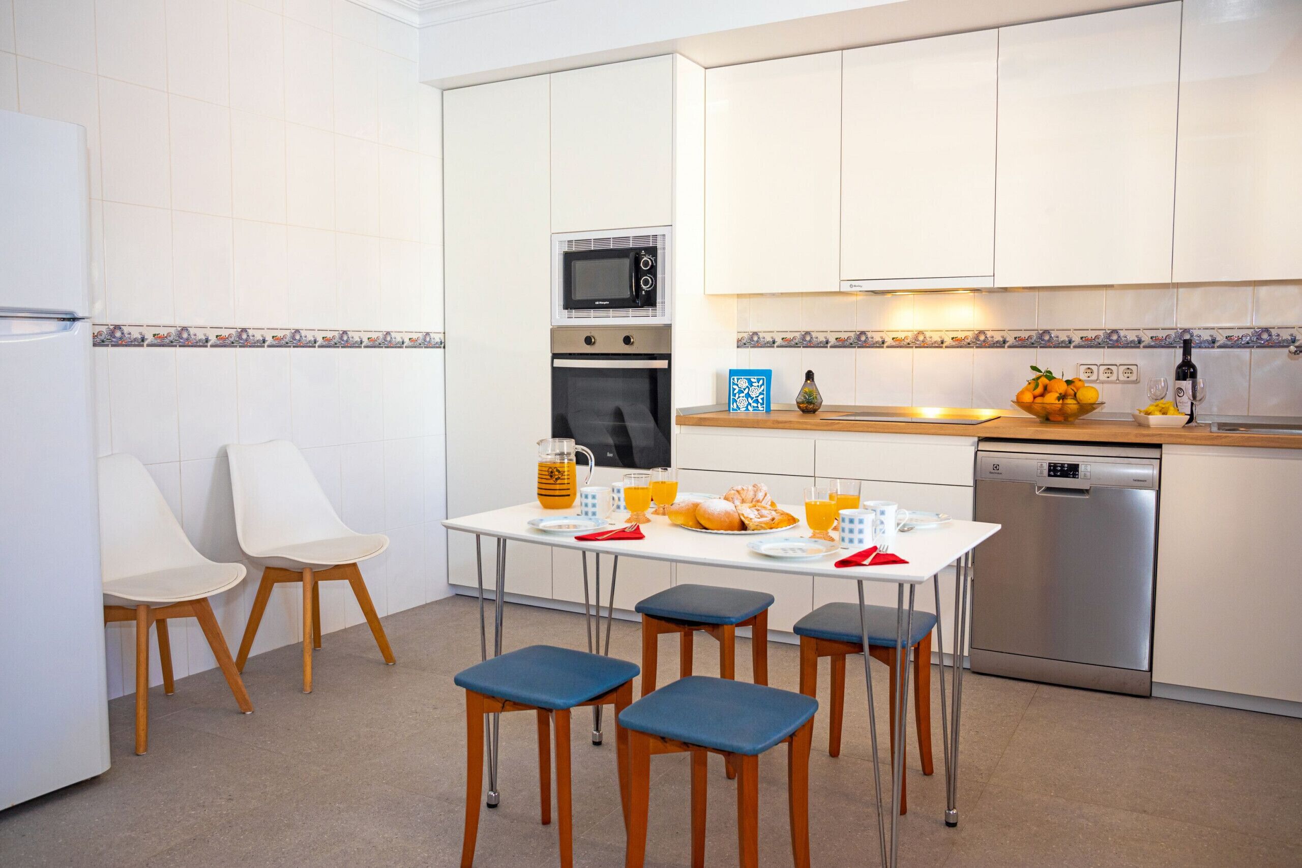 A modern kitchen with white cabinets, a microwave, oven, and dishwasher. There is a dining table with chairs and a fruit bowl.
