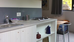 Une cuisine équipée d’un évier, d’une cuisinière et de divers ustensiles. Il y a une fenêtre avec des rideaux et une chaise dans le coin repas.