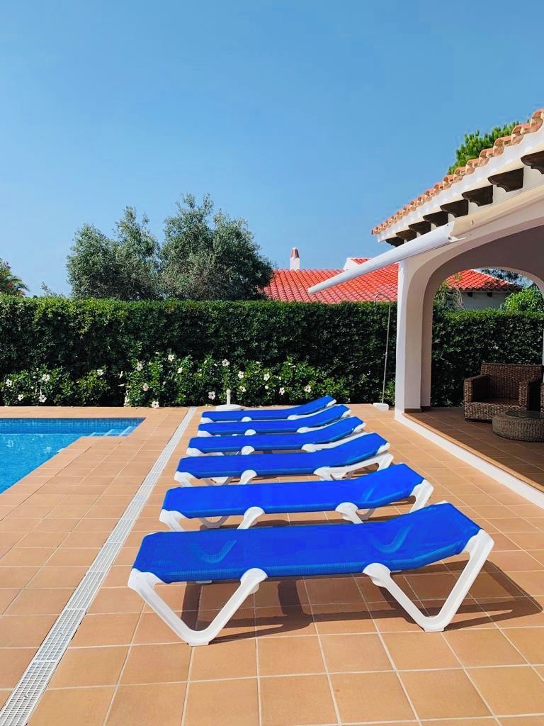 Un espace aménagé au bord de la piscine, avec des chaises longues bleues disposées en rangées, une terrasse carrelée et un coin salon couvert doté d’un toit recouvert de tuiles rouges.