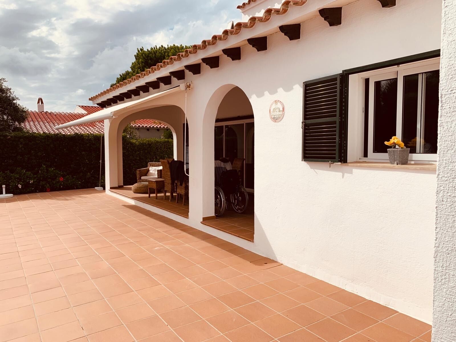 A covered patio area with a white wall and tiled floor, featuring a wheelchair-accessible entrance and a window with shutters.