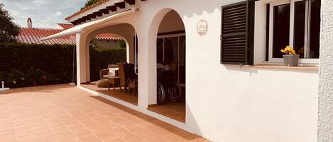 A covered patio area with a white wall and tiled floor, featuring a wheelchair-accessible entrance and a window with shutters.