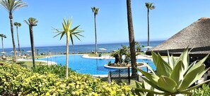 A pool area with a view of the ocean, surrounded by palm trees and a thatched-roof structure. - Mi Capricho 170 Beachfront Apartment (Mijas)
