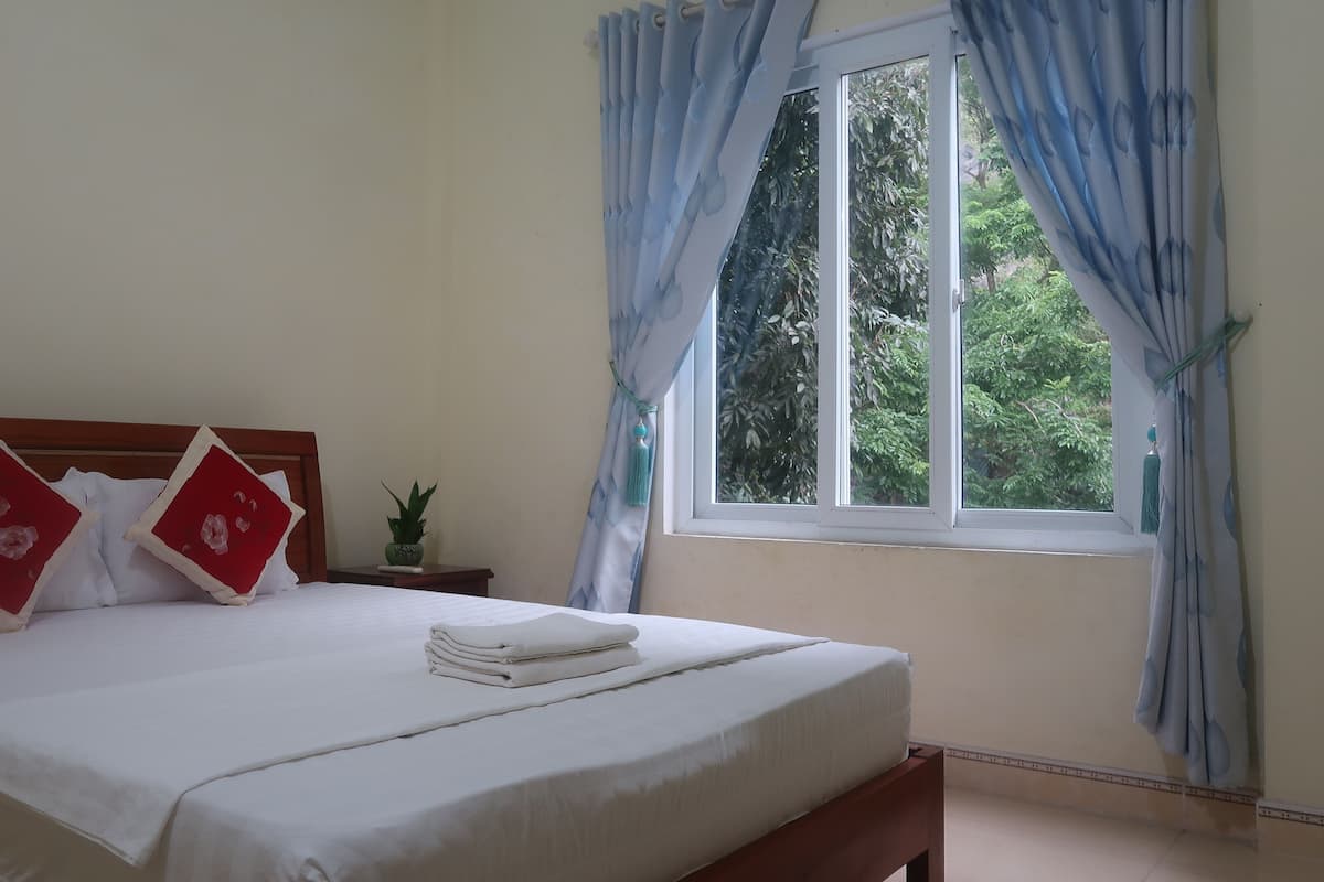 Una cama bien hecha con sábanas blancas y dos almohadas rojas, una mesita de noche con una planta y una ventana con cortinas azules.