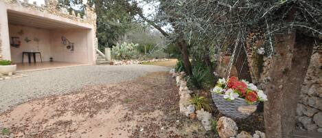 Un edificio rústico de piedra con una terraza abierta, un muro de piedra con una cesta de flores colgante y un camino de grava.