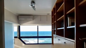 A room with a large window overlooking the sea, a ceiling fan, and built-in wooden shelving.