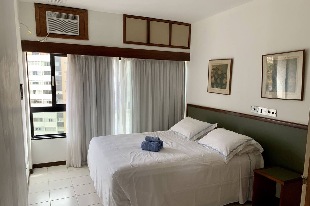 Une chambre d’hôtel avec un grand lit, une table de chevet, une fenêtre avec des rideaux et un climatiseur fixé au mur.