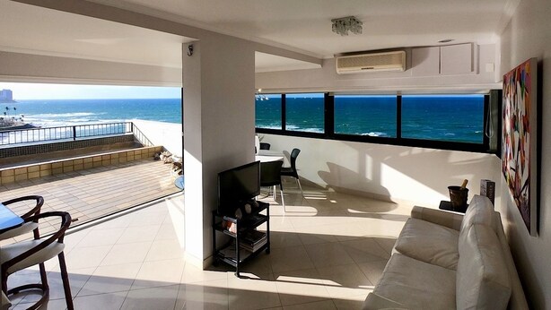A modern living room with a large window overlooking the ocean, a white sofa, a TV, and a dining area with chairs.