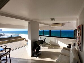 A modern living room with a large window overlooking the ocean, a white sofa, a TV, and a dining area with chairs.