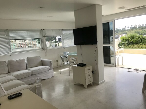 Un moderno salón con sofá, televisor montado en la pared y balcón con vistas a edificios y zonas verdes.