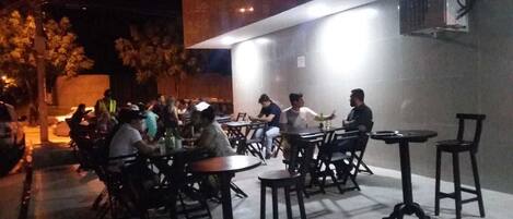 An outdoor seating area with tables and chairs, surrounded by a low wall and trees.