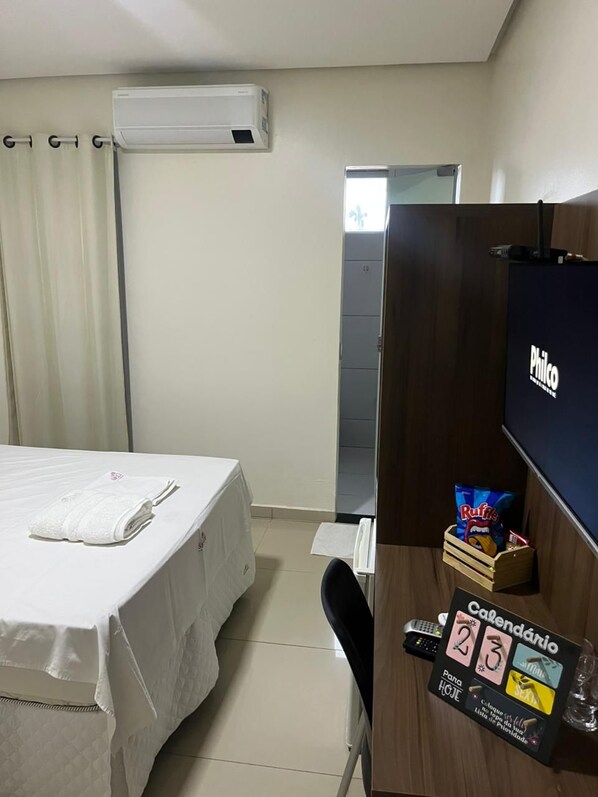 A hotel room with a bed, a desk with a calendar, a TV, and a small wooden box with snacks.