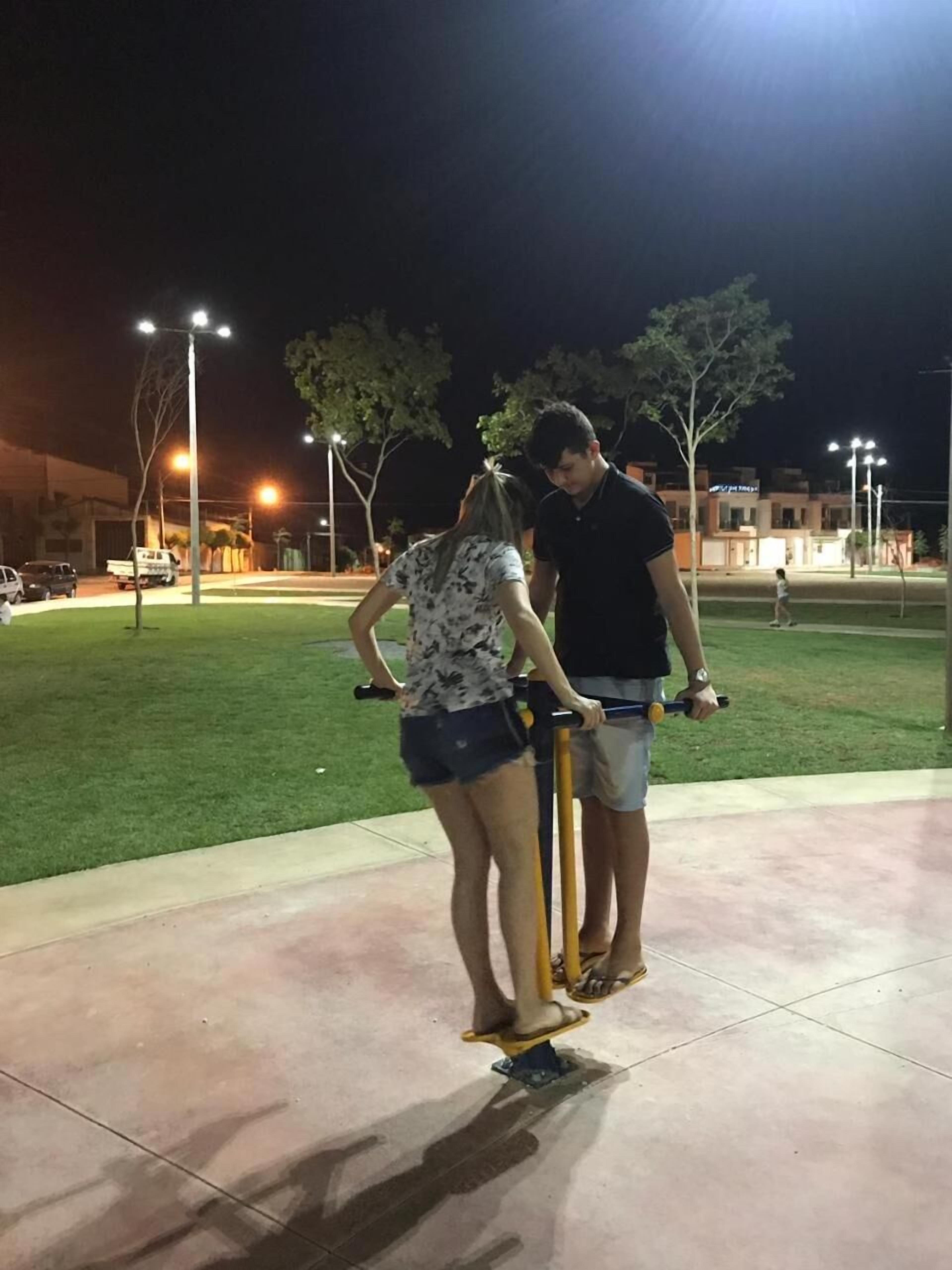 Two people on a scooter in an outdoor area with a paved path, trees, and buildings in the background.
