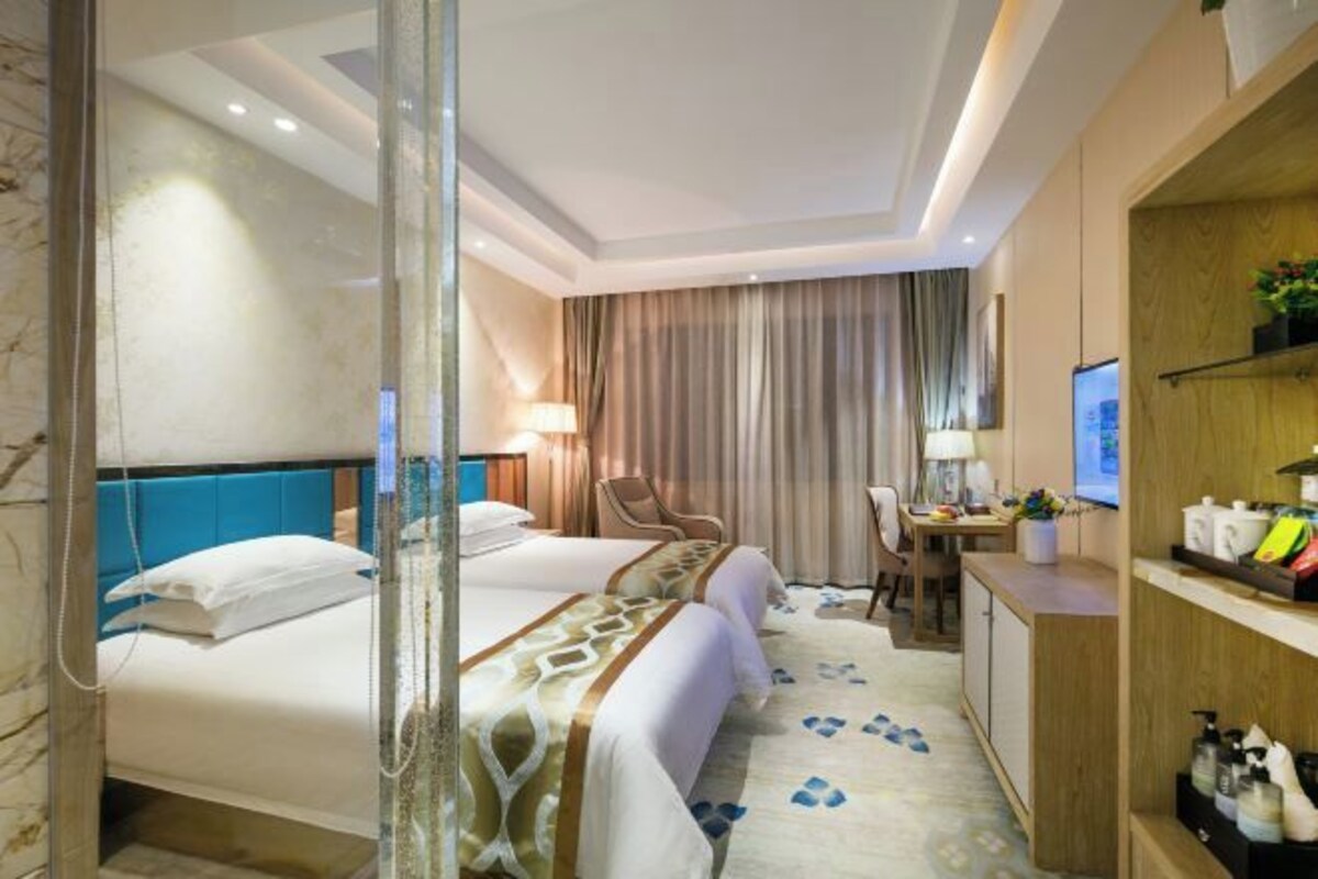 A hotel room with two beds, a desk, a chair, a TV, and a bathroom with a glass shower.