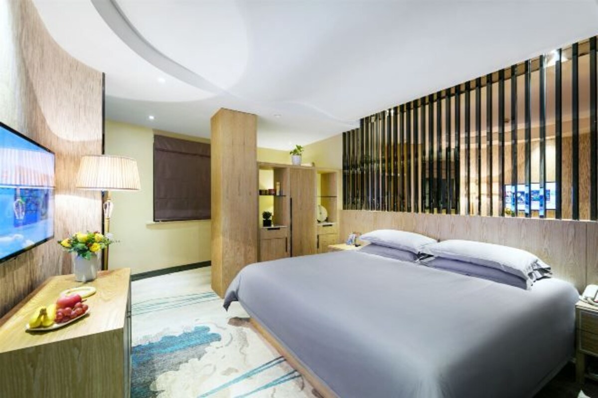 A modern hotel room with a large bed, a desk with a lamp, a TV, and a decorative wall panel.
