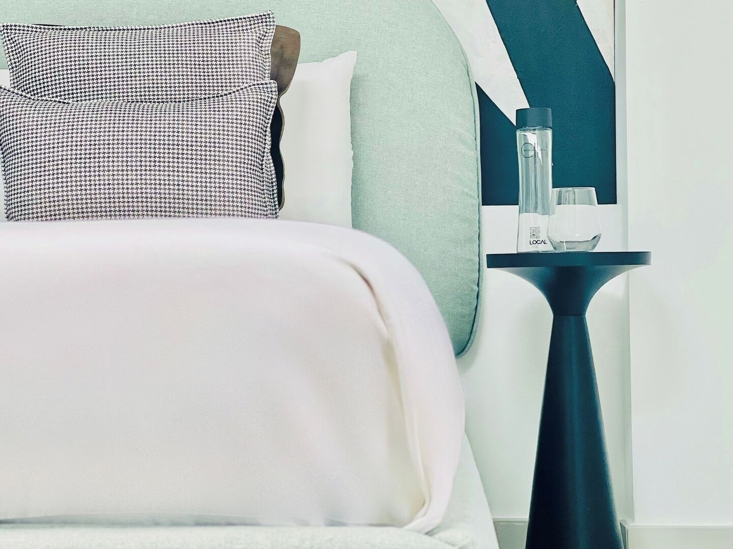 A neatly made bed with a white comforter and a black and white patterned pillow, a small table with a glass and a bottle, and a wall with a geometric design.