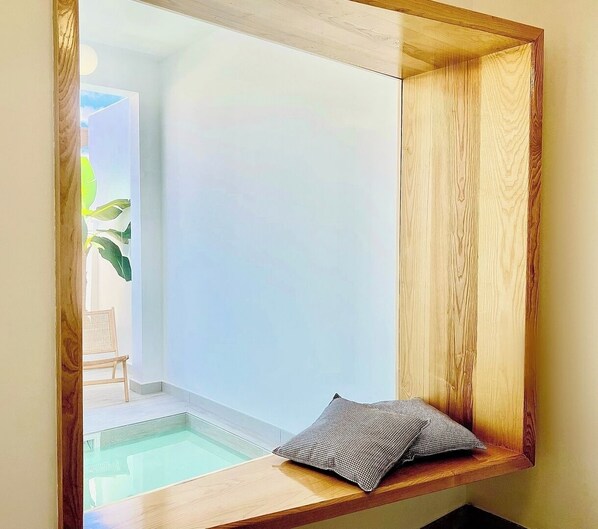 A corner of a room with a wooden bench and a cushion, a small pool, and a view of a plant and a chair in the background.