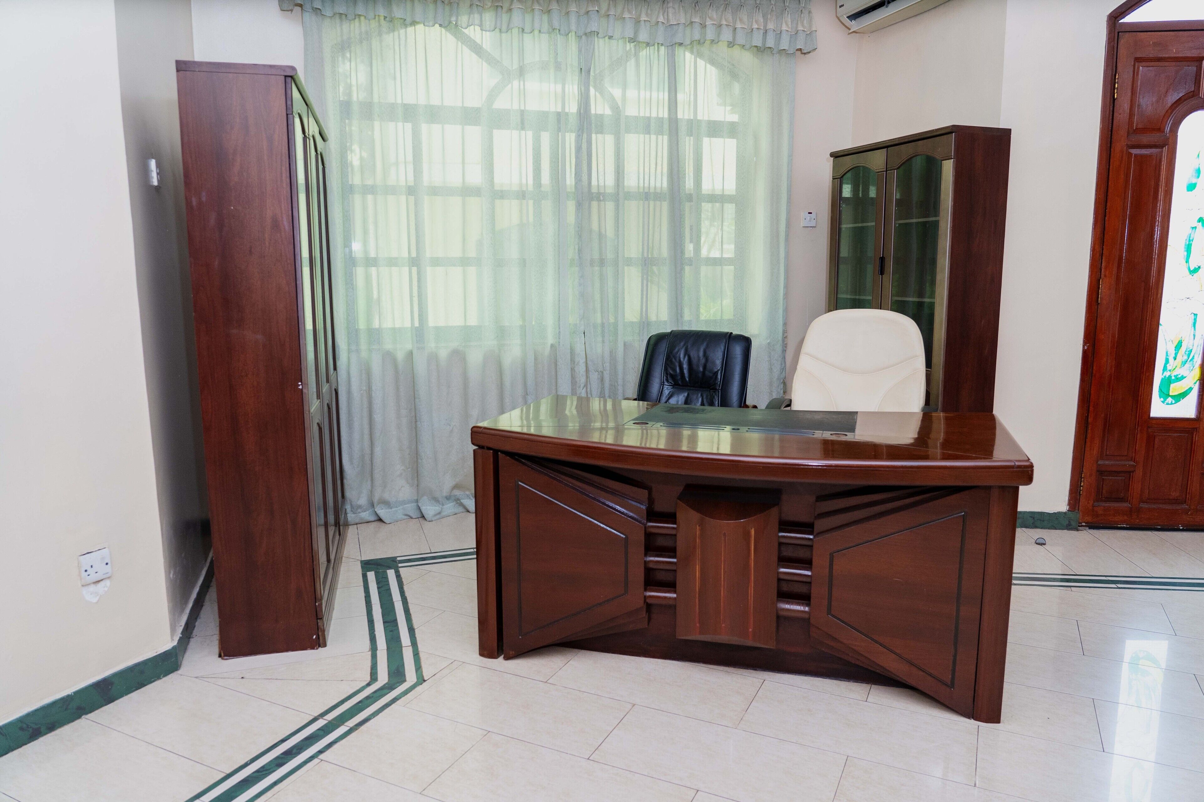 A wooden desk with drawers, a leather chair, and a glass cabinet in a room with a tiled floor and a window with sheer curtains.