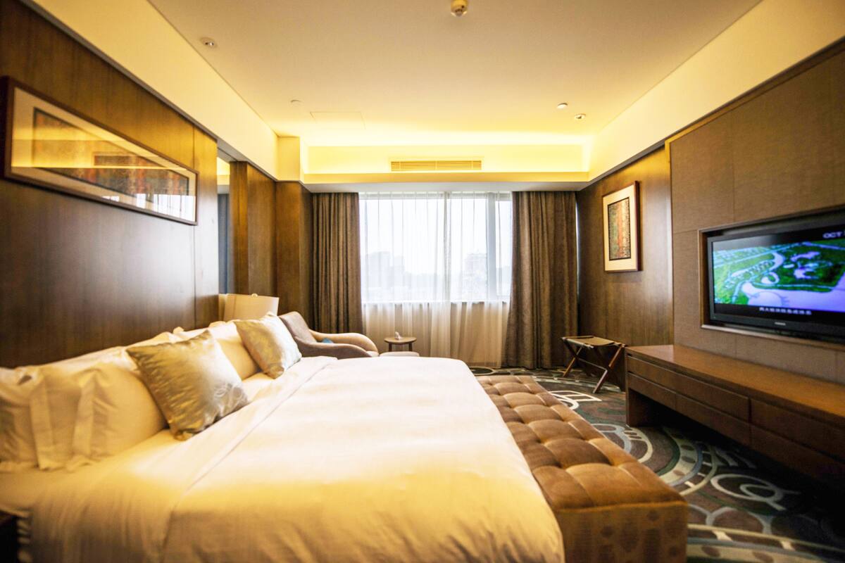 A hotel room with a large bed, a TV, a seating area, and a view of the city through the window.
