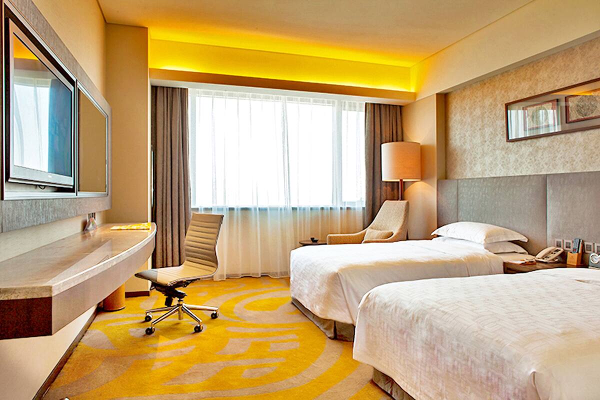 A hotel room with two beds, a desk with a chair, a desk lamp, and a large window with curtains.