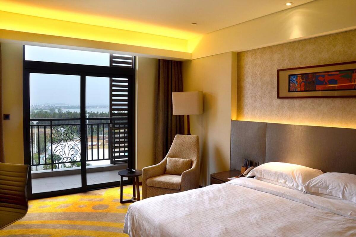 A hotel room with a large bed, a chair, a nightstand, a lamp, and a view of the outdoors through a balcony.