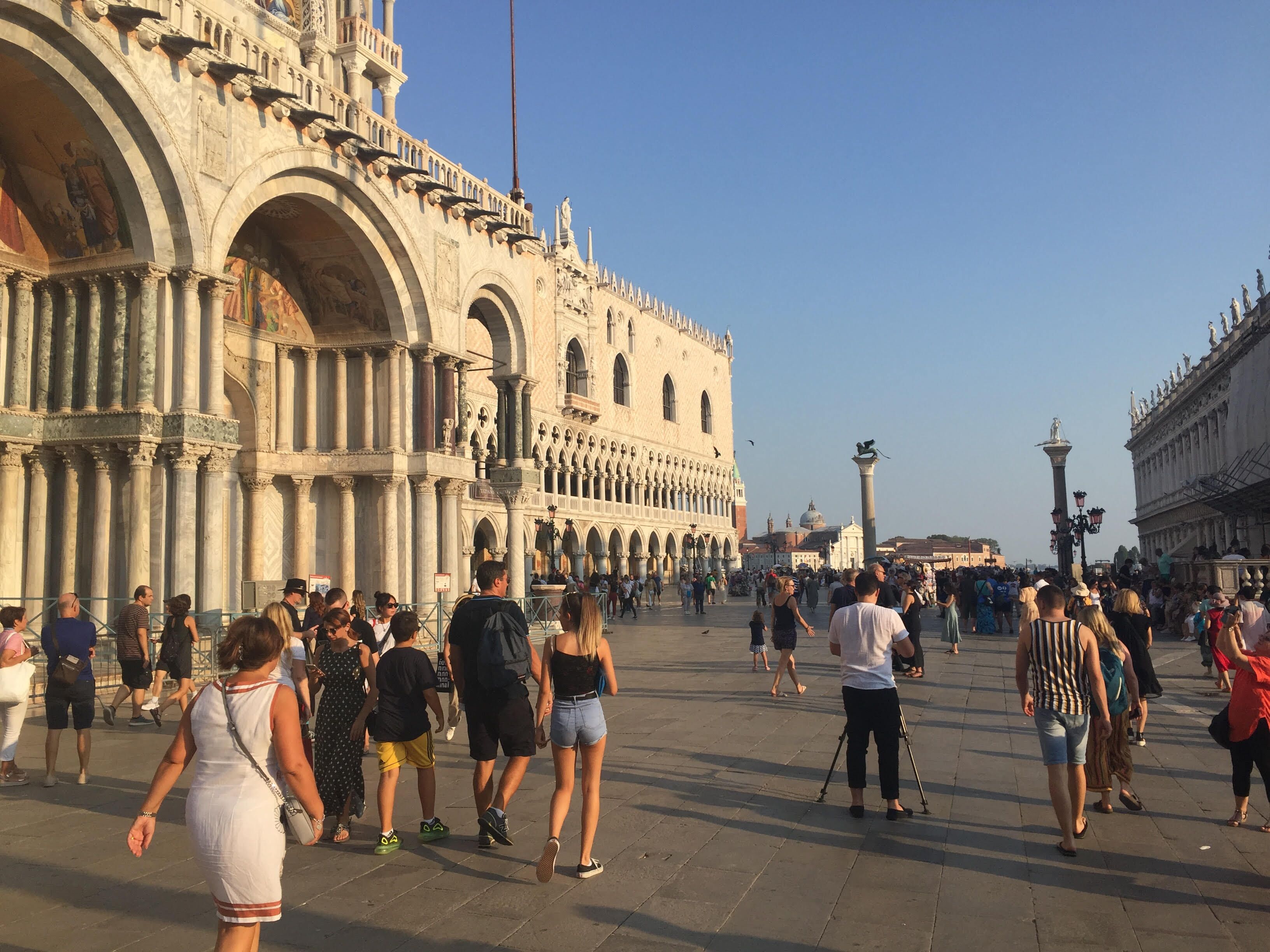 A historic building with arched windows and columns, a large open plaza, and people walking around.