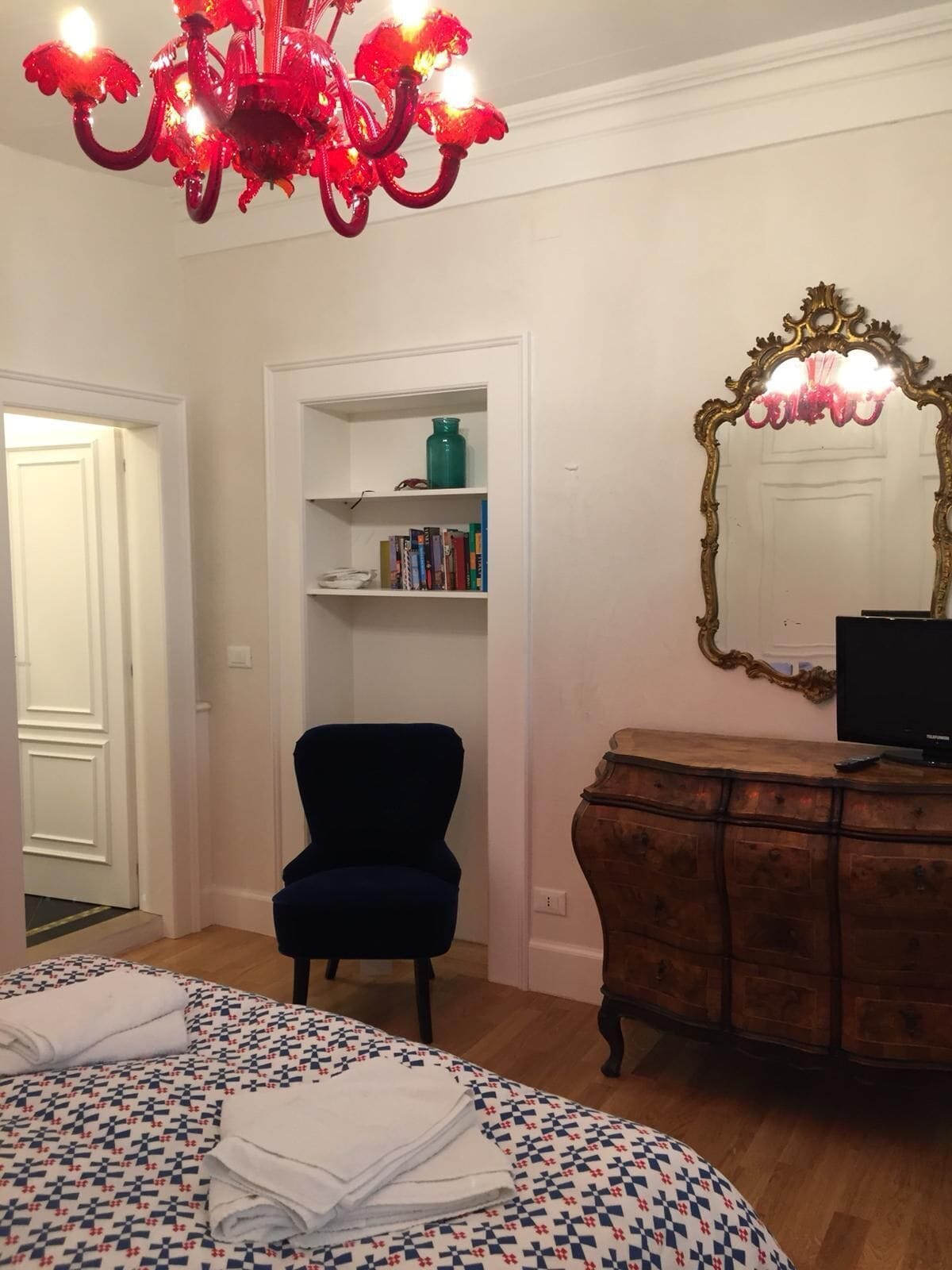 A room with a bed, a blue armchair, a wooden dresser, a bookshelf, and a mirror.