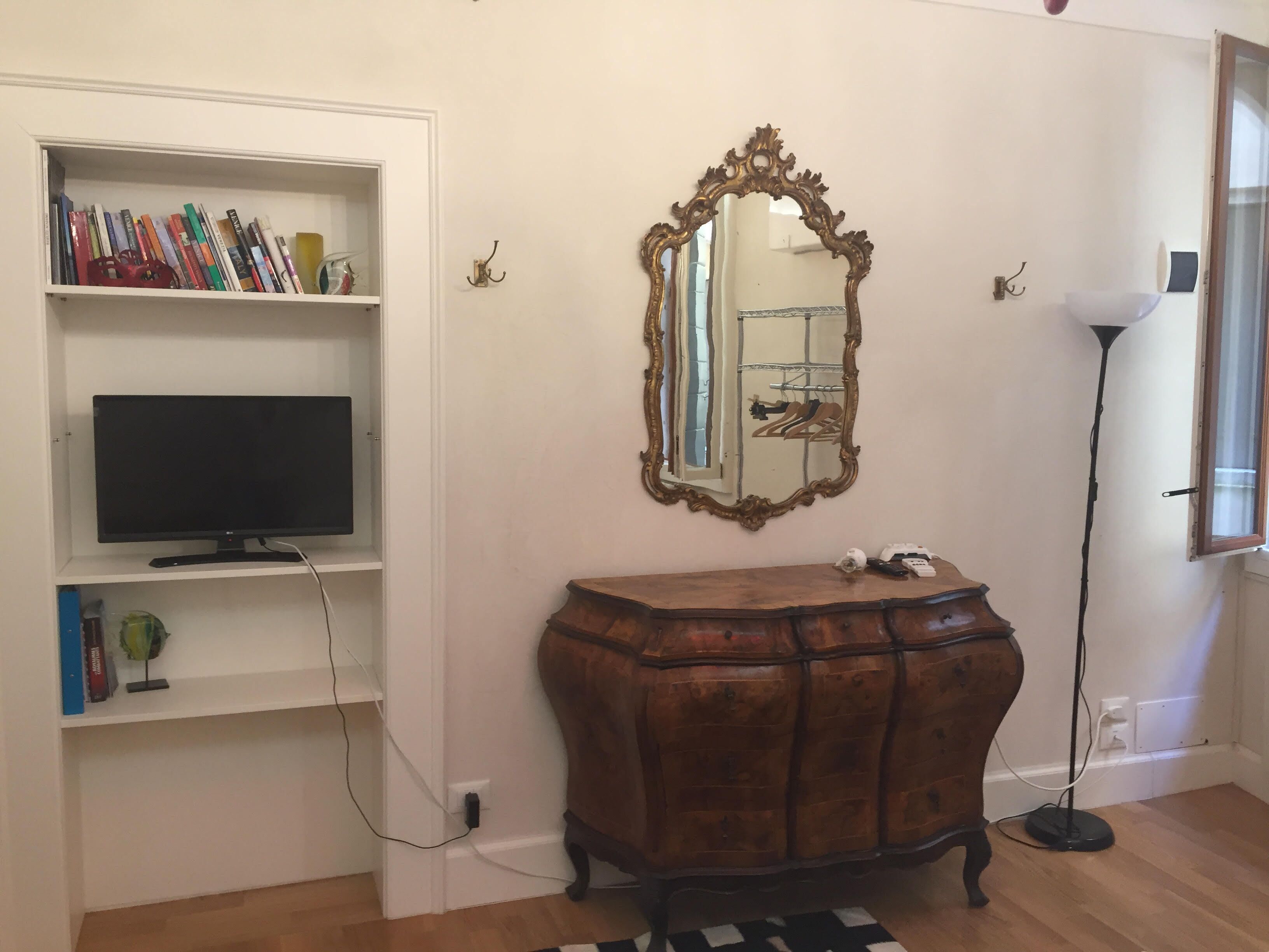 Una habitación con pared blanca, cómoda de madera, televisor en un estante, espejo y lámpara de pie.