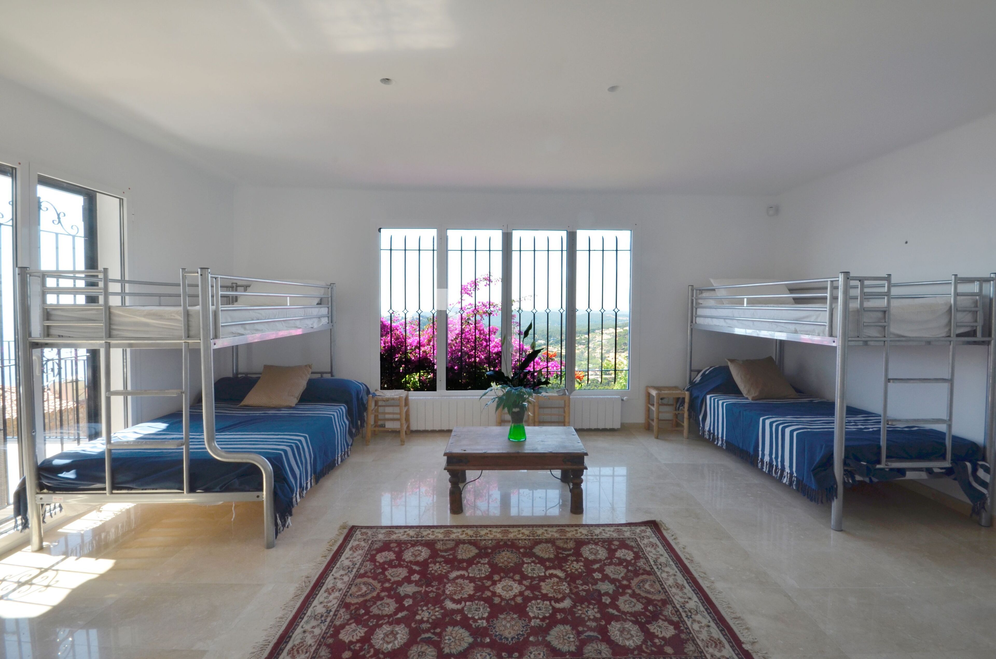 A room with bunk beds, a central table, and a large window with pink flowers.