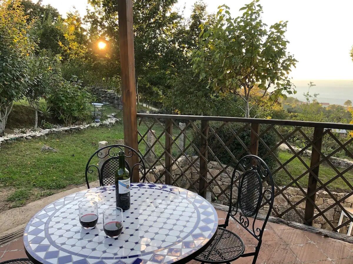 Une table ronde recouverte d’une nappe à motifs, deux verres de vin rouge et une bouteille de vin, le tout sur une terrasse offrant une vue sur la mer.