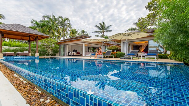 A swimming pool with blue mosaic tiles, surrounded by lounge chairs and umbrellas, with palm trees and a covered seating area in the background. - Baan Ban Buri (Koh Samui)