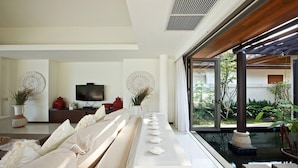 A modern living room with a long white sofa, a flat-screen TV, decorative wall art, and a view of a garden through large windows. - Baan Ban Buri (Koh Samui)