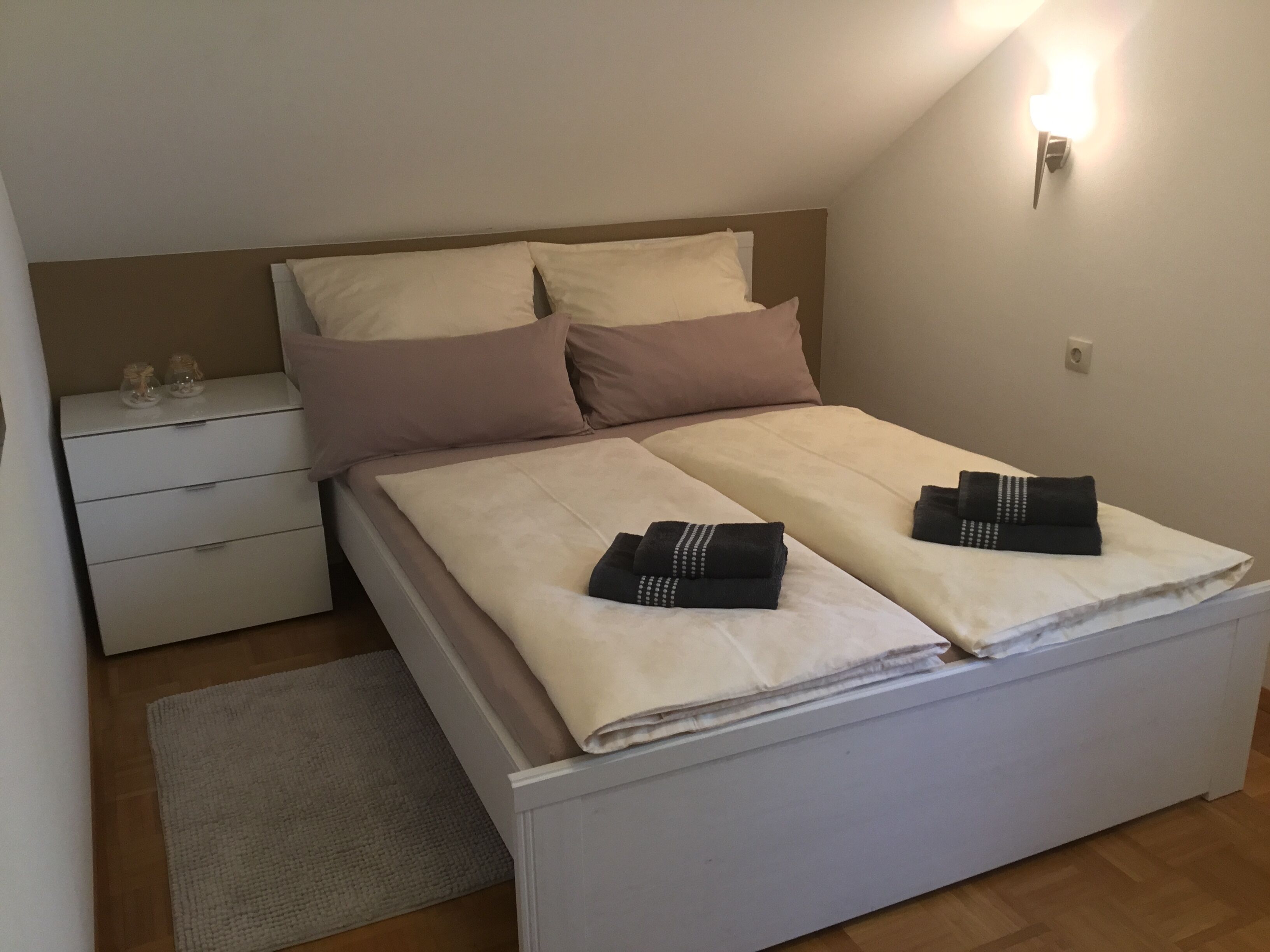 A neatly made bed with two pillows, a bedside table with glasses, and a wall-mounted light.