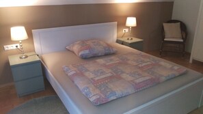 Een net opgemaakt bed met een bedsprei met patroon, twee bedlampjes, een stoel en een nachtkastje met een lamp.