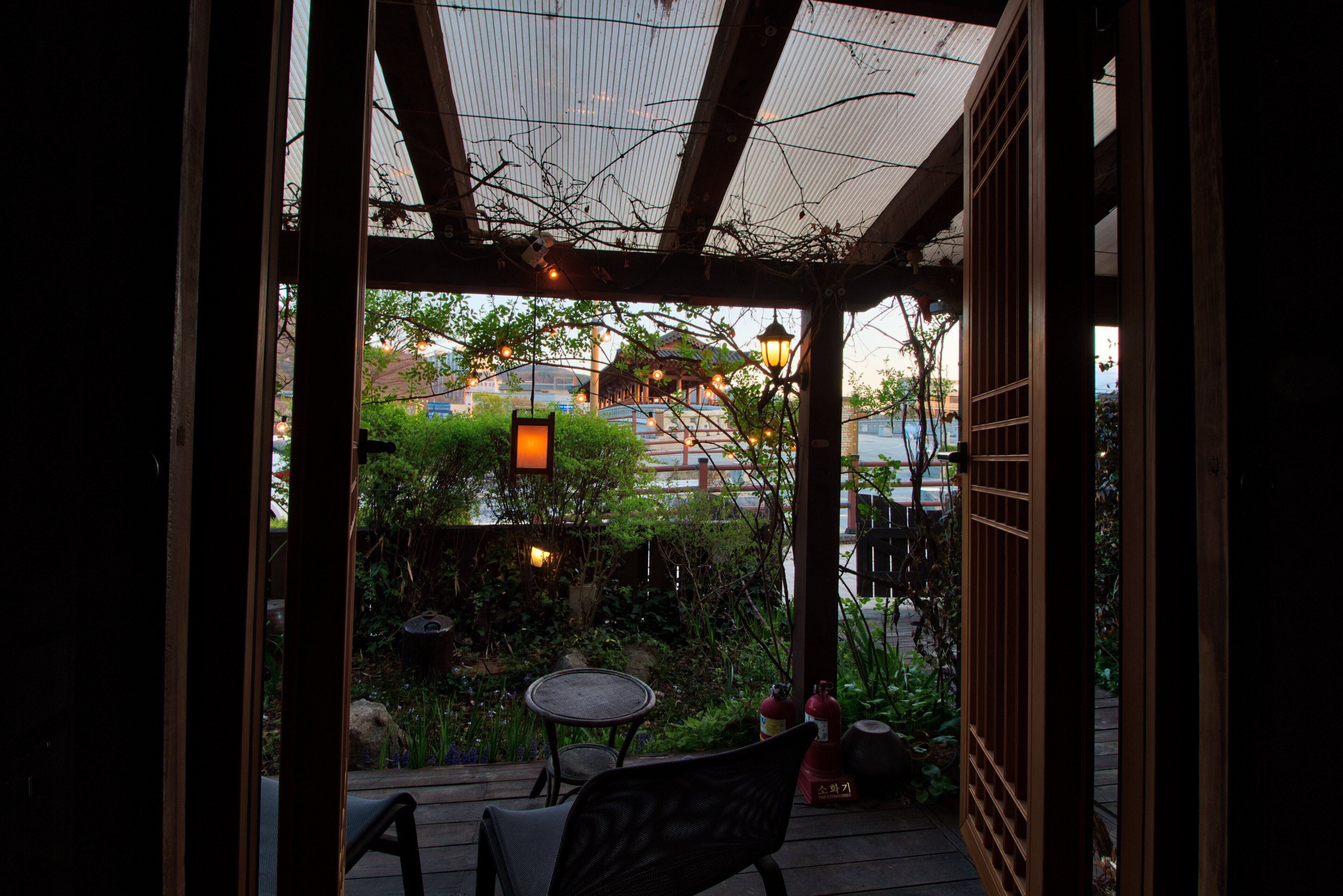 An outdoor patio with a wooden table and chairs, surrounded by greenery and a view of a building in the background.