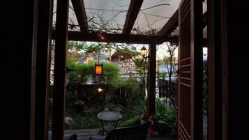An outdoor patio with a wooden table and chairs, surrounded by greenery and a view of a building in the background.