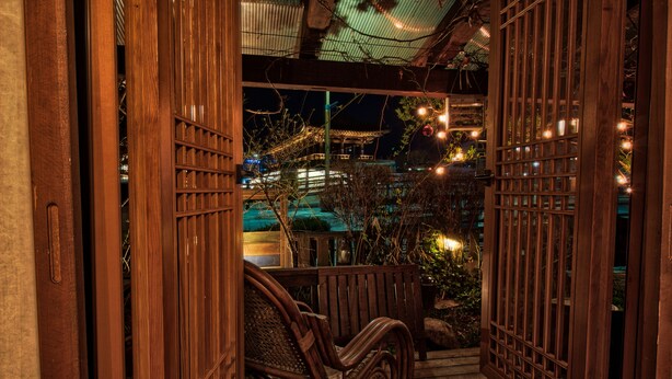 Une porte en bois mène à un porche où se trouve un fauteuil en osier. On peut admirer un bâtiment illuminé et un jardin.