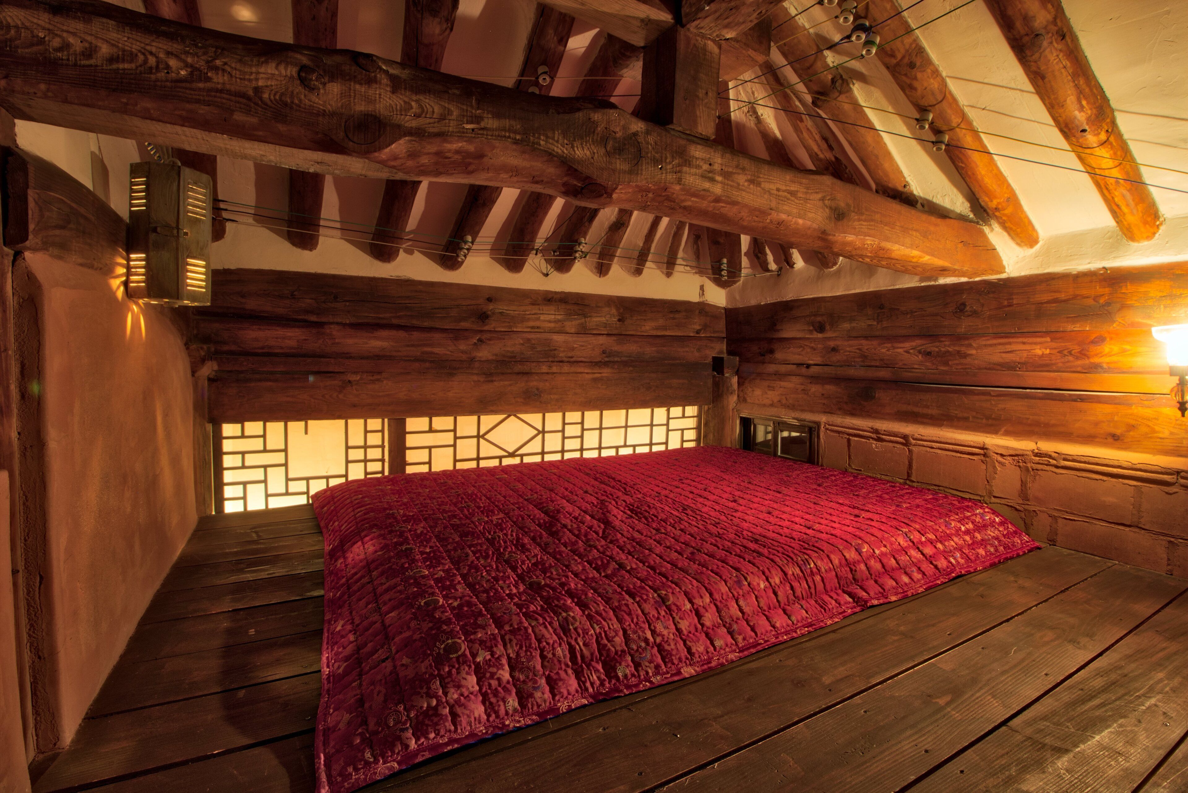 A wooden cabin with a sloped ceiling, a bed with a red blanket, and a window with a geometric pattern.