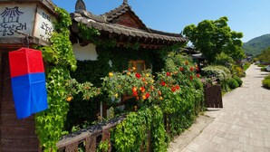 Une maison traditionnelle coréenne, avec un toit recouvert de tuiles, entourée d’une végétation luxuriante et de plantes en fleurs.