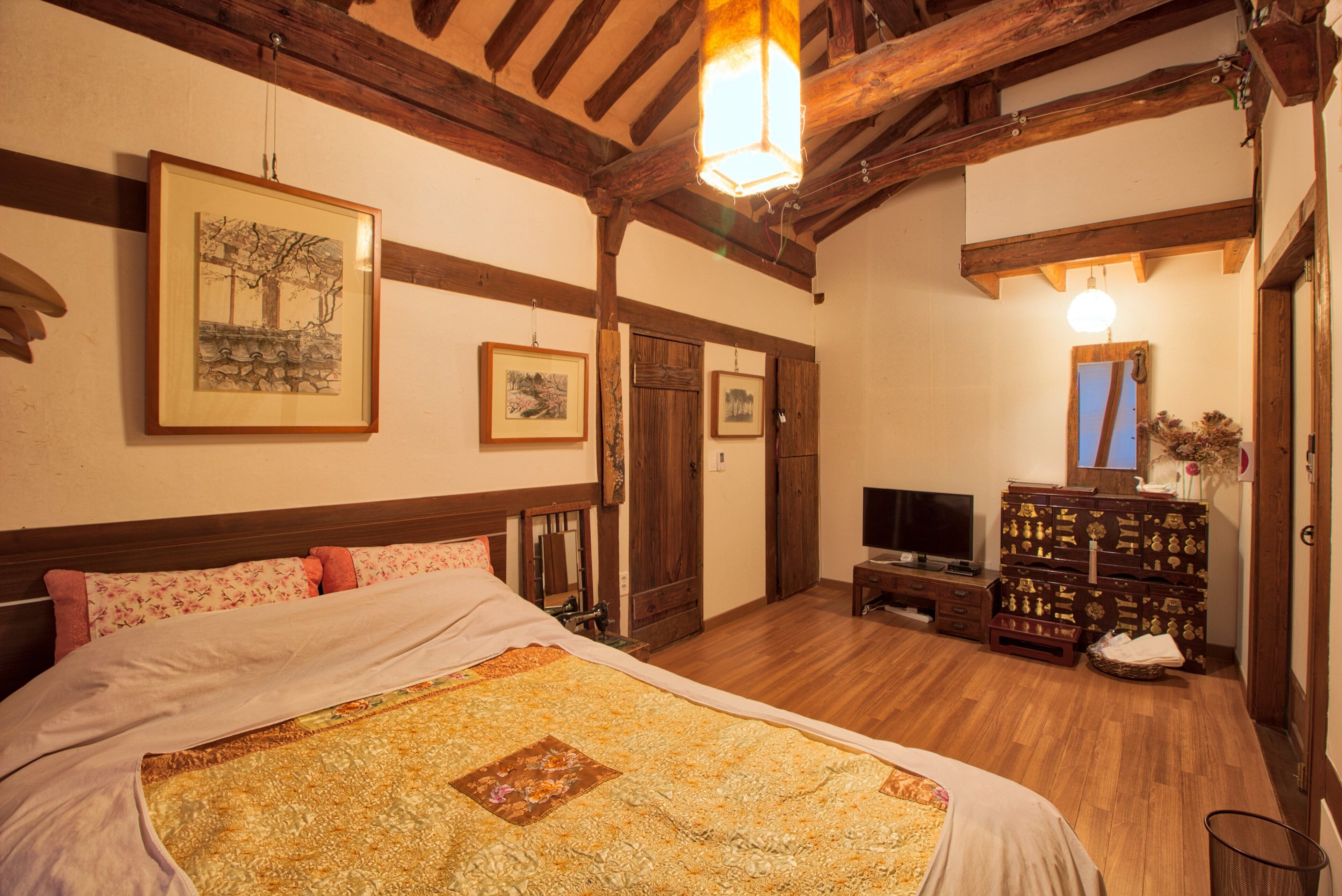 A bedroom with a large bed, wooden beams, framed artwork on the walls, a television, and a dresser with drawers.