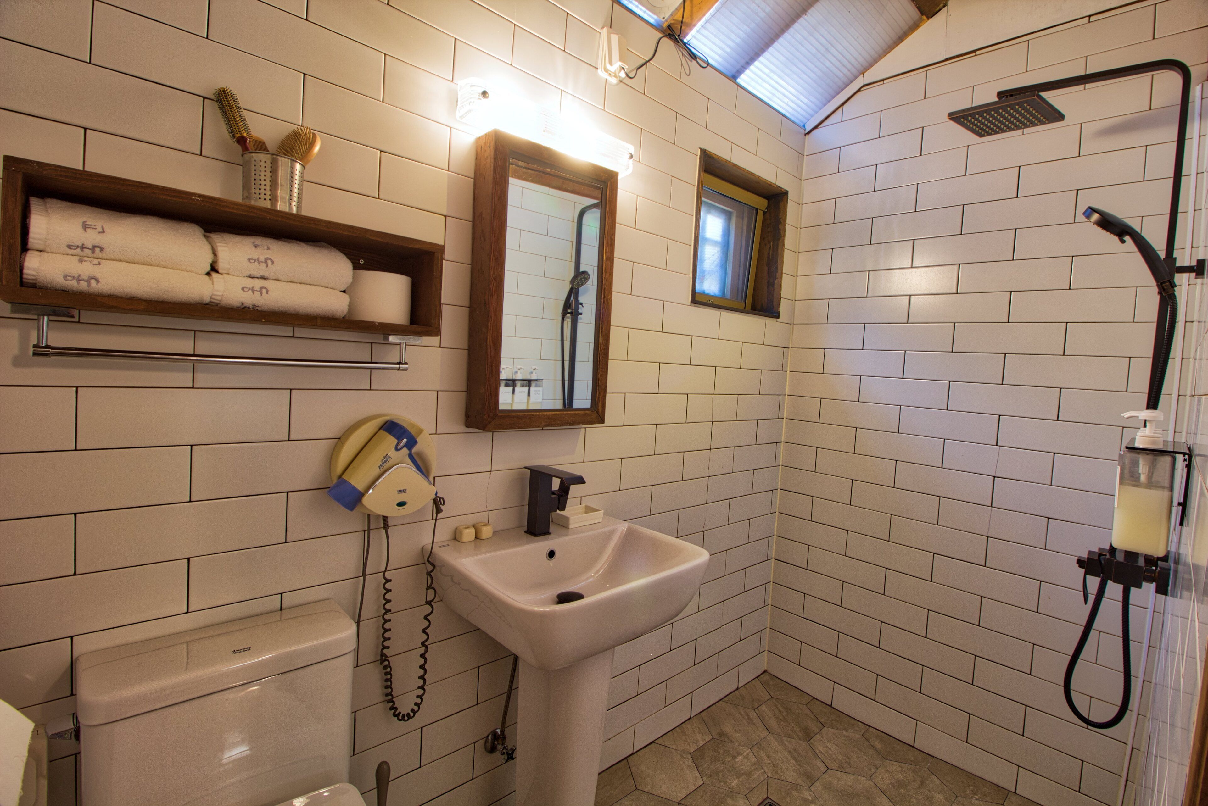 Baño con azulejos blancos tipo metro, lavamanos blanco, espejo, ducha y teléfono.