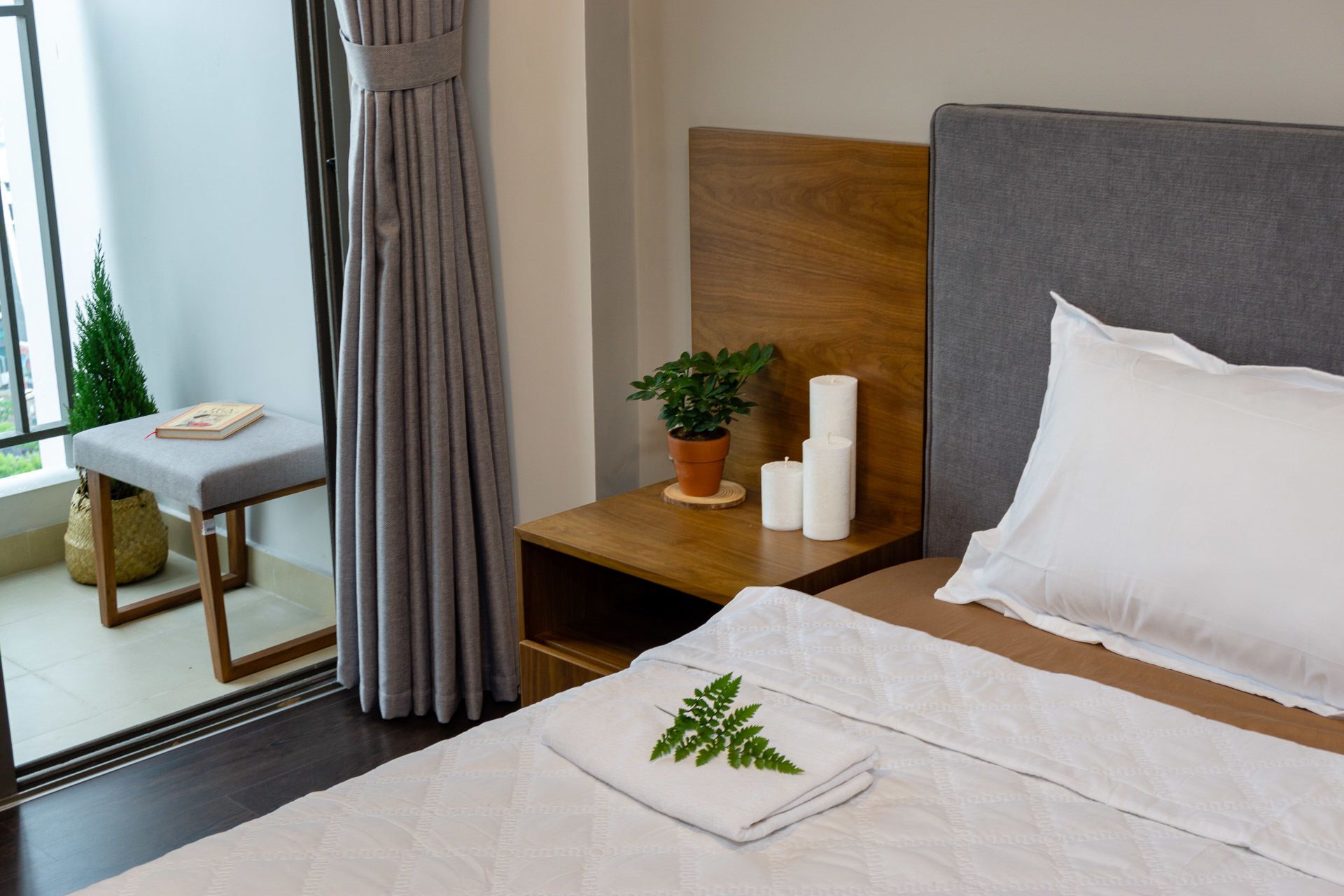 Une chambre d’hôtel bien rangée, avec un lit, une table de chevet ornée de bougies, une petite plante en pot et une fenêtre avec des rideaux.