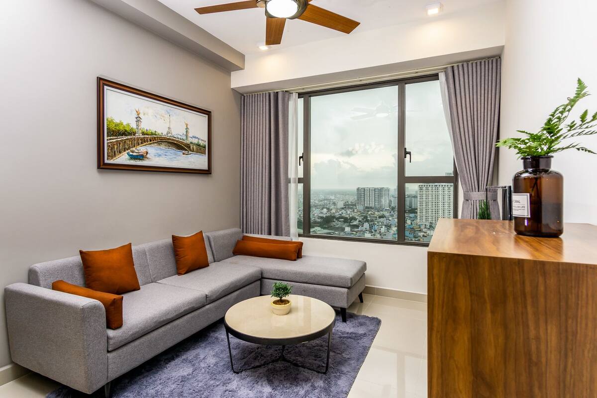 Uma sala de estar moderna com um sofá seccional cinza, uma mesa de centro redonda e vista para a cidade através de grandes janelas.